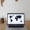 Laptop with black world map on screen placed on wooden table near white wall with silhouettes of continents